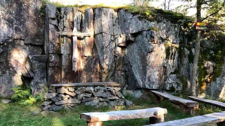 Metsäkirkko ulkona, kallioinen seinä, jossa risti, edessä puisia penkkejä