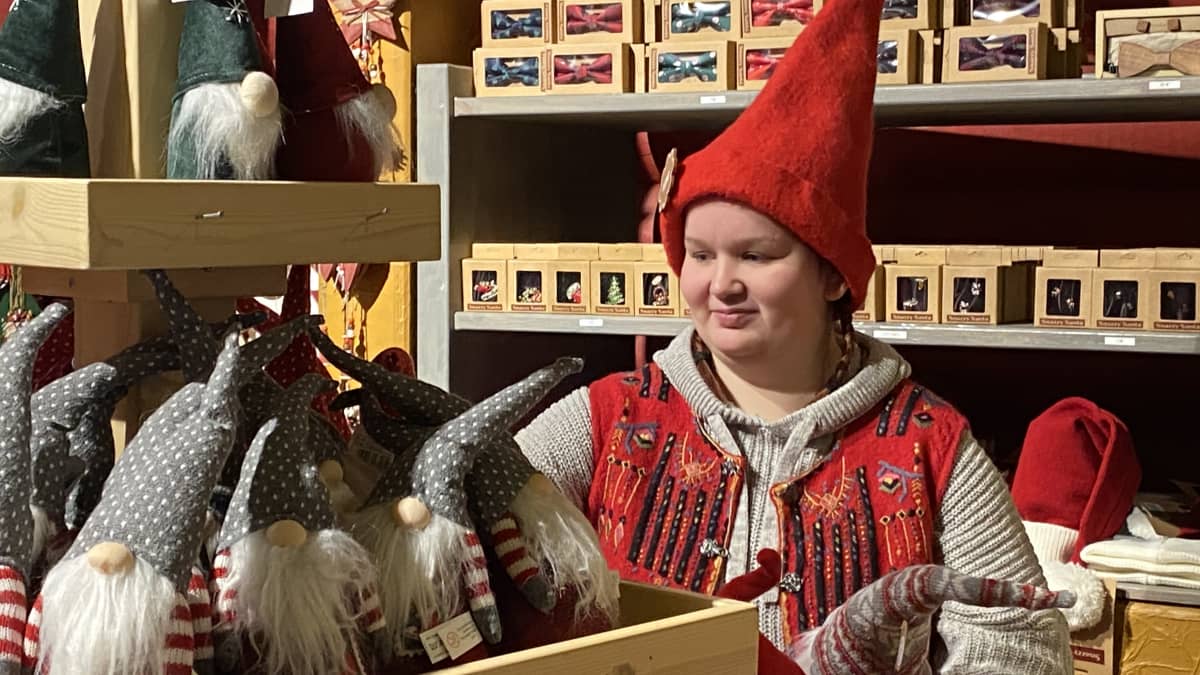 Punaiseen piippalakkiin pukeutunut tonttu-asiakaspalvelija järjestää pehmo-tonttuja myymälän hyllyyn.