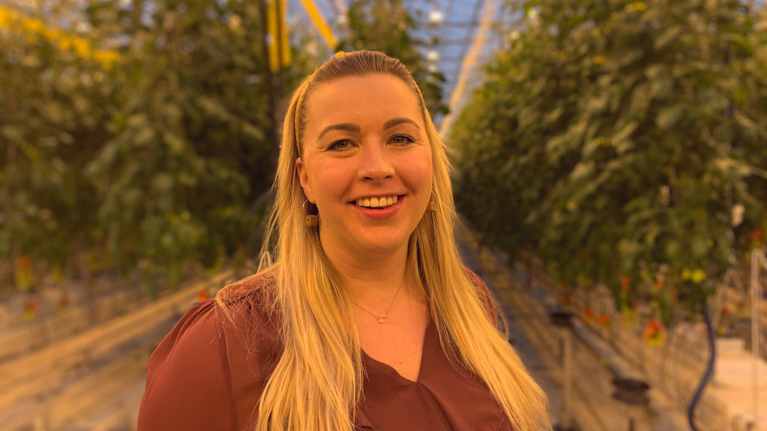 Kvinna med långt ljust hår ler mot kameran. I bakgrunden växthusmiljö med tomatplantor.