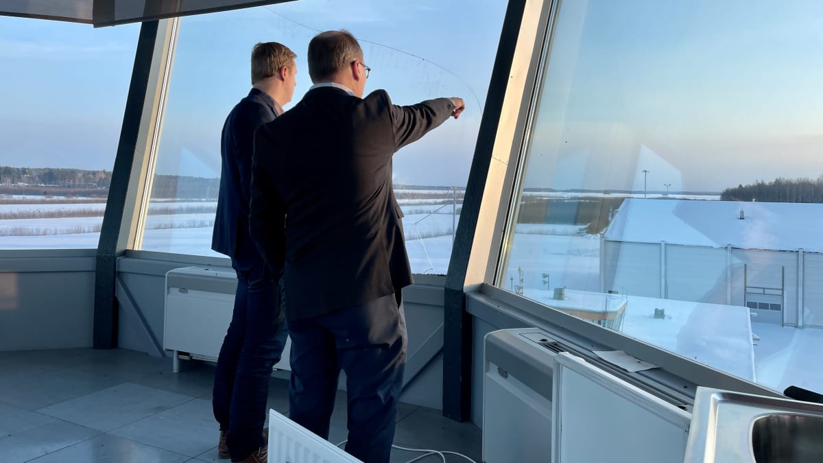 Kaksi miestä seisoo korkeassa lennonjohtotornissa ja katsoo aurinkoista maisemaa.