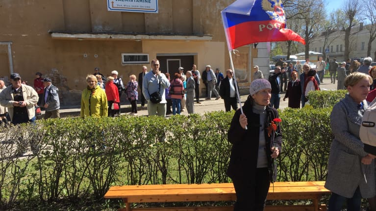 Mustiin vaatteisiin pukeutunut vanhempi nainen pitää punaisia neilikoita ja heiluttaa Venäjän lippua aurinkoisena kevätpäivänä virolaisessa Kohtla-Järven kaupungissa. 
