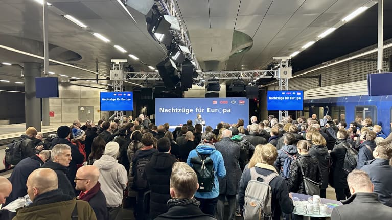 Ihmisjoukko seisoo selin kameraan ja katsoo lavalle, jossa lukee "Yöjunia Eurooppaan" saksaksi. Vasemmalla juna.