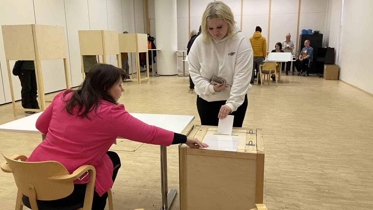 Nainen istuu selin pöydän ääressä pinkki jakku päällä ja auttaa kasvot kameraan päin olevaa vaaleaan huppariin pukeutunutta naista pudottamaan äänestyslipukkeen uurnaan.