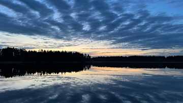 Rauhallinen järvimaisema auringonlaskun aikaan, jossa sinertävä taivas ja pilvet heijastuvat veden pinnasta.