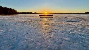 Aamu Littoistenjärvellä. Aurinko nousee horisontista, puistonpenkki keskellä jääkenttää. 