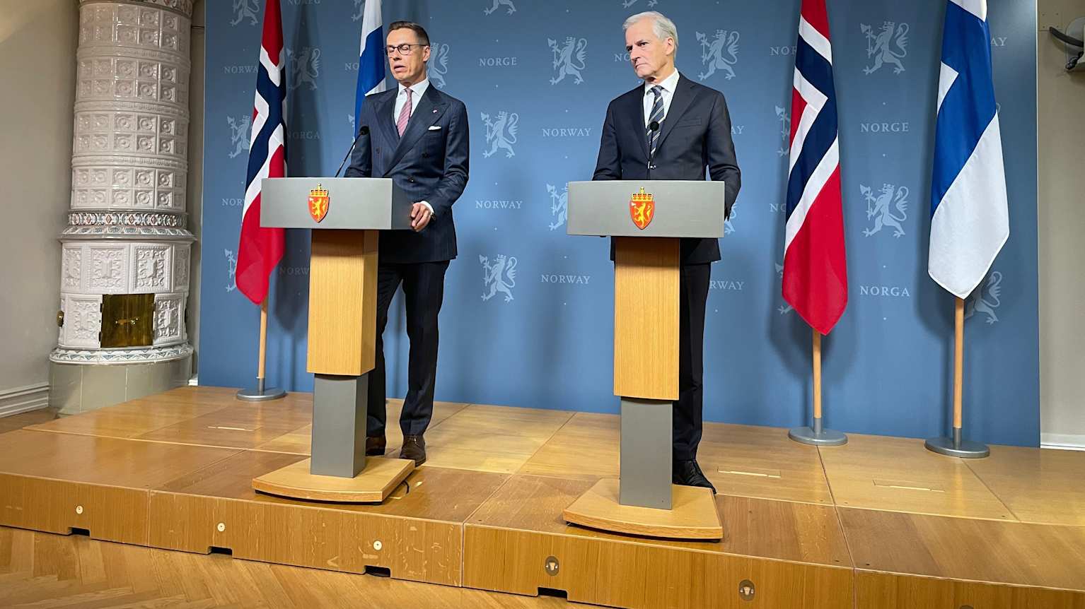 Two men in dark blue suits standing behind podiums with 4 Norwegian and Finnish flags and an old-fashioned tiled heating stove behind them.