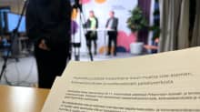 Etualalla on paperi, jossa kerrotaan aluehallituksen päättävän maanantaina sote-asemien, kiirevastaanottojen ja vuodeosastojen palveluverkosta. Taustalla on epätarkasti ihmisiä ja kameroita.