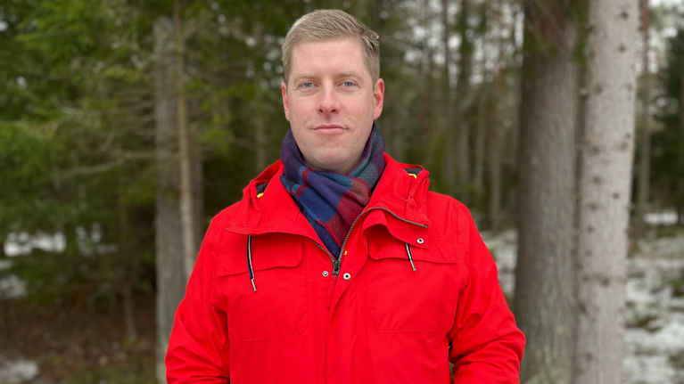 Man med röd jacka står framför en skog och tittar in i kameran.