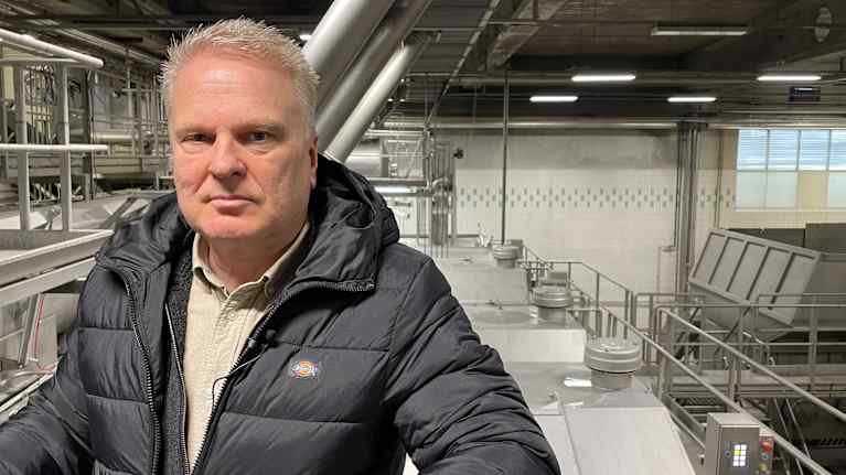 En man i svart täckjacka står uppe på en ställning framför stora aluminumtankar i en industrihall. 