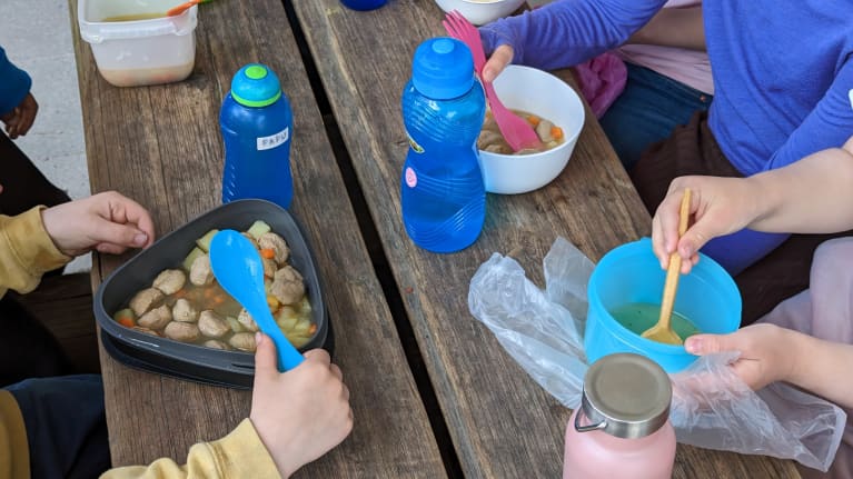 Pöydällä erilaisia muoviastioita ja aterimia joilla lapset syövät siskonmakkarakeittoa.