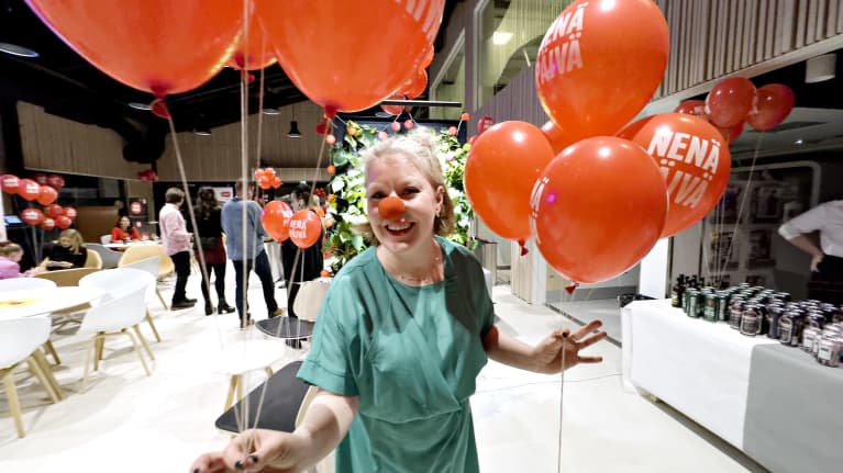 Vihreään mekkoon pukeutunut, vaaleahiuksinen nainen (toimittaja Julia Falck) hymyilee leveästi kameraan katsoen punainen Nenäpäivä-nenä päässään ja punaisia ilmapalloja käsissään. 