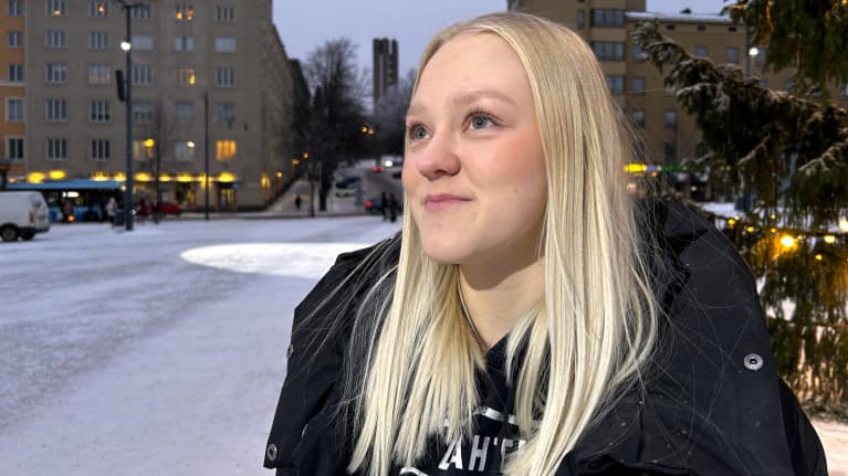 Vaaleatukkainen nuori nainen seisoo joulukuusen vieressä ja katsoo taivaalle. 
