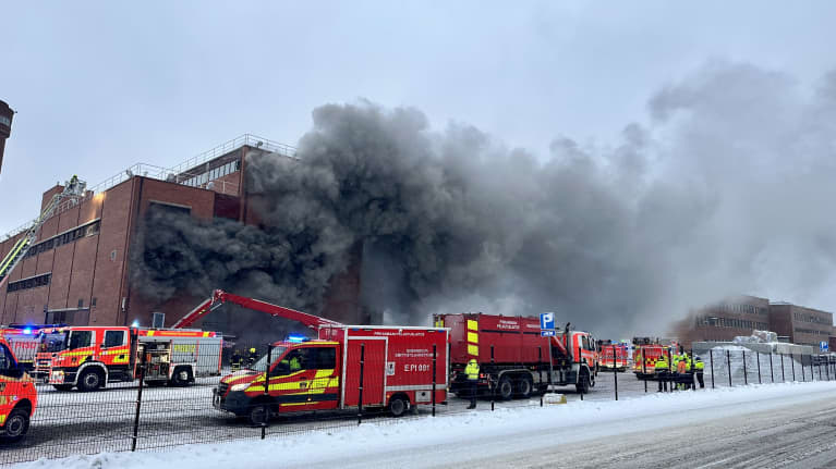 Tulipalo Nokian paperitehtaalla. Kuvassa punatiilisestä rakennuksesta nousee paksua mustaa savua, pelastushenkilökuntaa korkean nosturin päällä. Kuvauspäivä 29.11.2023.
