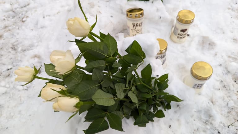 Valkoisia ruusuja ja surukynttilöitä lumessa tien reunassa.