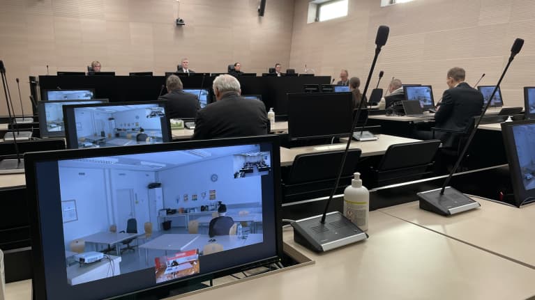Oikeussalissa istuu edessä tuomarit ja yleisössä asianajajat. Syytetyt näkyvät tietokoneen ruudulla.