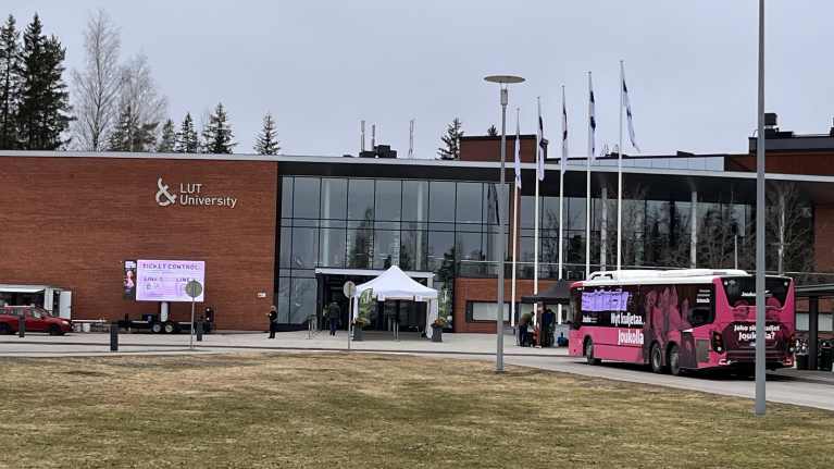 Pinkki bussi seisoo punatiilisen yliopistorakennuksen ulkopuolella.