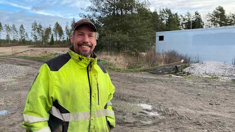 Glad medelålders jordbrukare står stort leende i arbetskläder och keps intill en lerig åker.