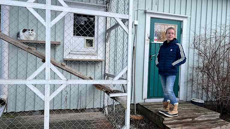 Siniseen toppatakkiin pukeutunut henkilö seisoo rakennuksen portailta. Verkkoaitauksen sisäpuolella henkilön vieressä pötköttää kissa.
