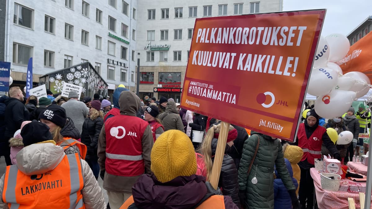 Takaapäin kuvattuna ihmisiä seisomassa rotuaarin aukiolla. 