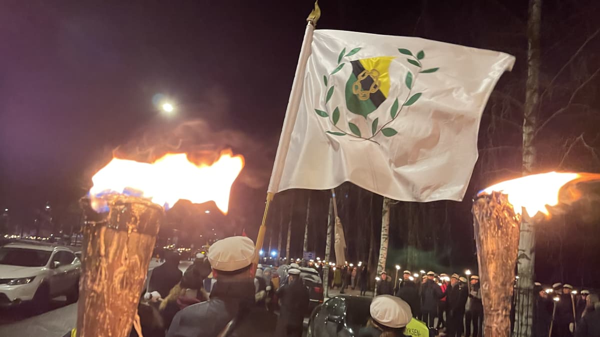 Människor i studentmössor går i fackeltåg den 6.12.2021. En del håller i facklor, en i en stor vit flagga.