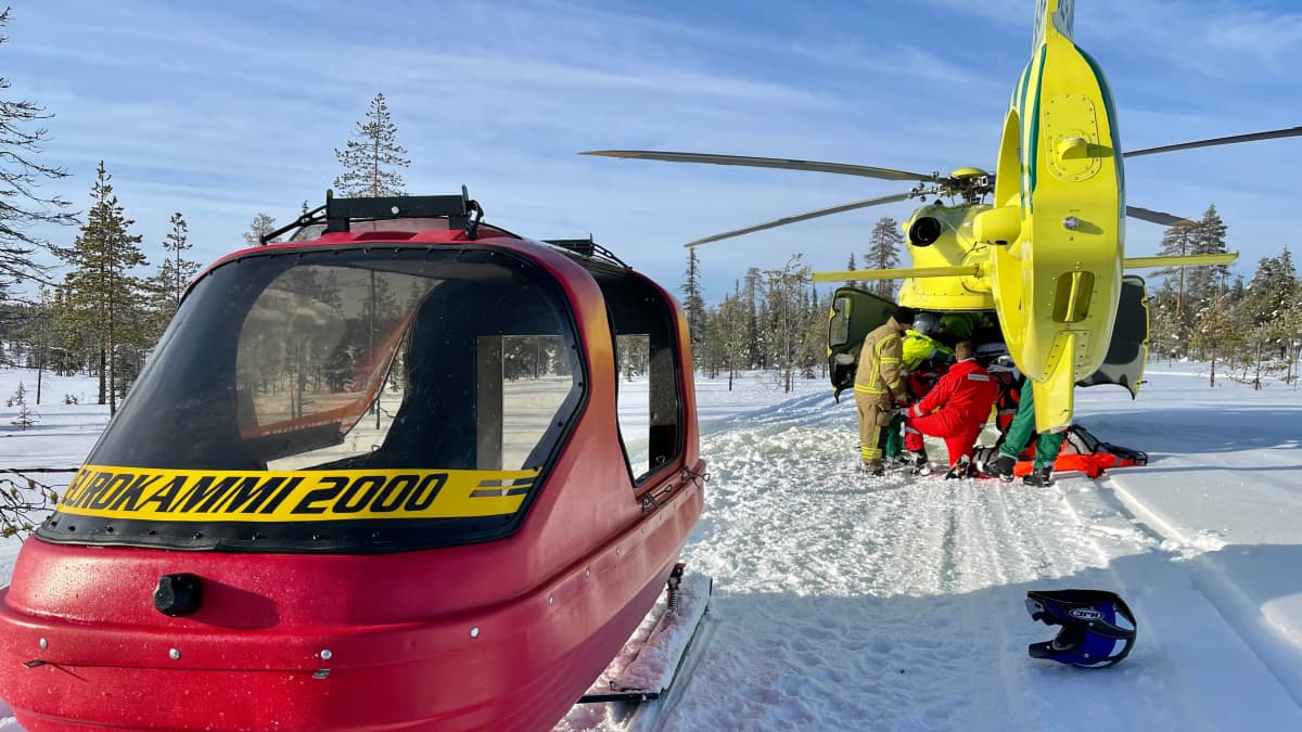 Punainen moottorikelkan perässä vedettävä pelastusreki hangella kelkkareitillä. Taustalla kirkkaankeltainen pelastushelikopteri.