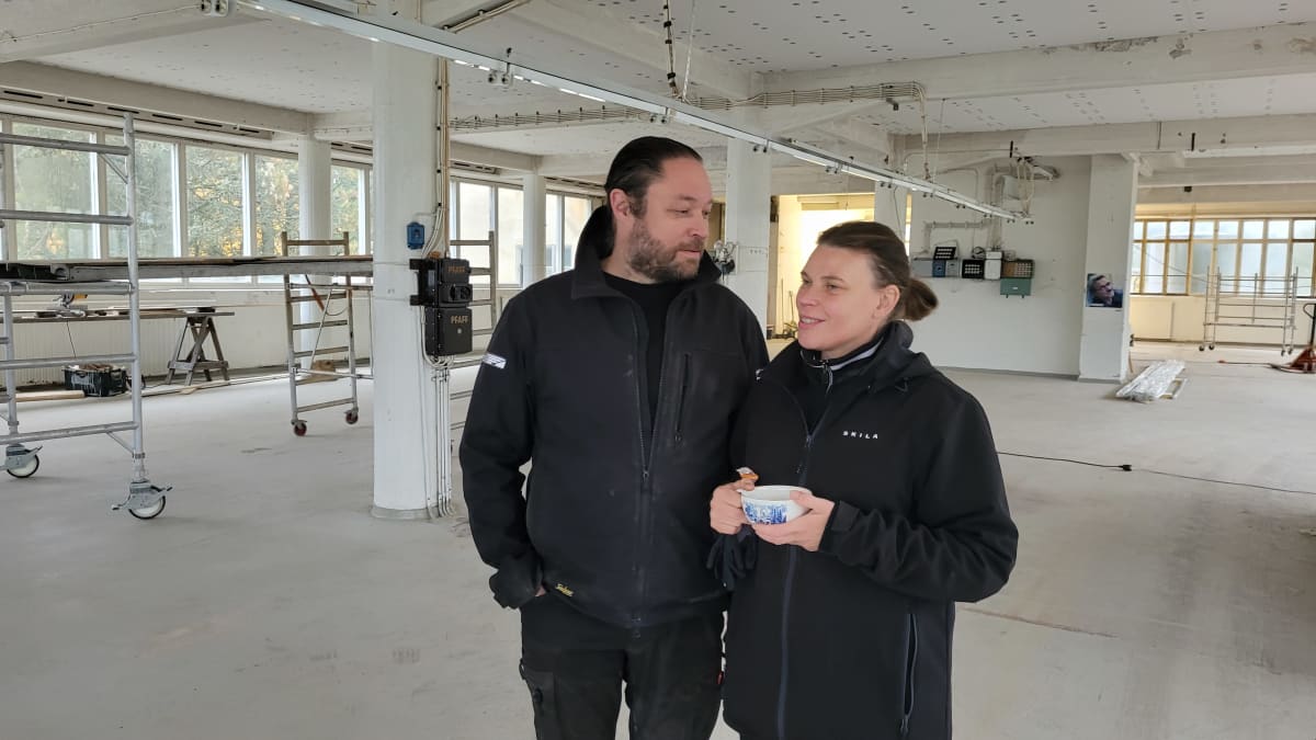 Juha Ollila ja Päivi Hakkarainen seisovat keskellä valkoista betonista hallitilaa. Taustalla rakennustelineitä. Nainen pitelee kädessään sinivalkoista teekuppia.