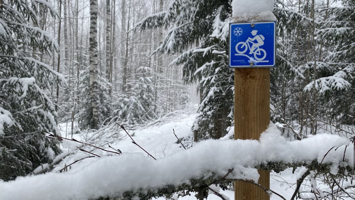 Lumisessa metsässä olevassa puupalkkiin kiinnitetyssä kyltissä on valkoinen pyöräilijähahmo sinisellä taustalla. 