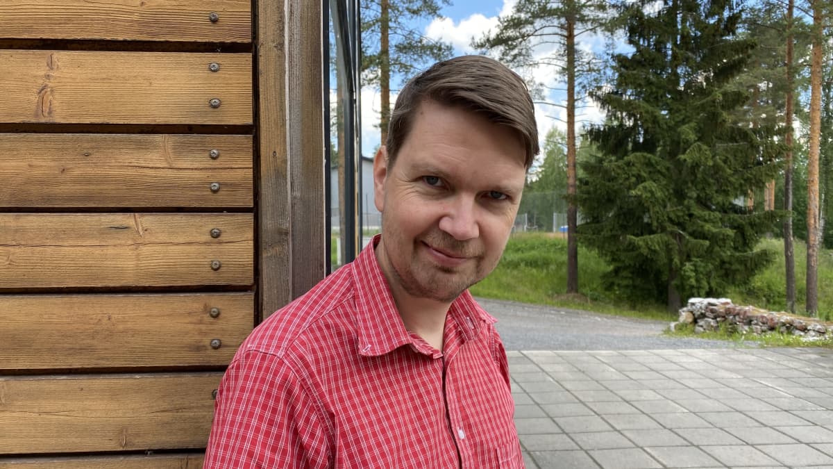 Matti Hakamäki nojaa Kansantaiteenkeskuksen nurkkaan etupihalla, jossa taustalla näkyy kuusi ja urheilualueita