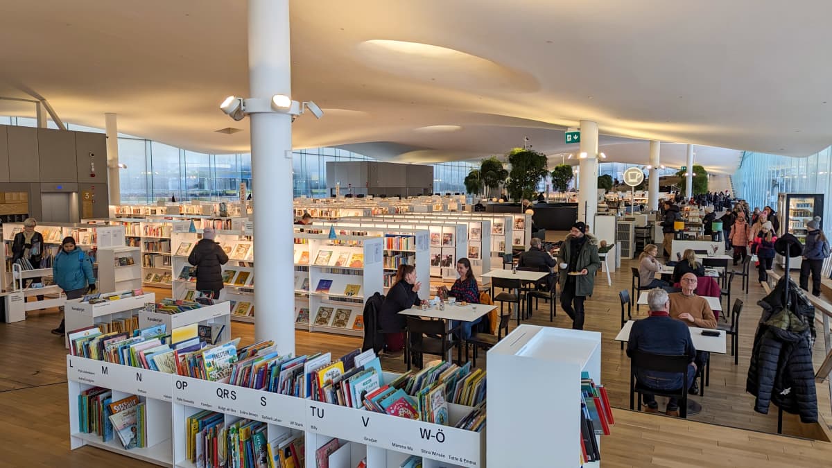 오드 도서관 최상층에 위치한 넓은 공간. 오른쪽에는 사람들이 있는 카페의 테이블과 의자가 있고 왼쪽에는 흰색 책장이 줄지어 있습니다.
