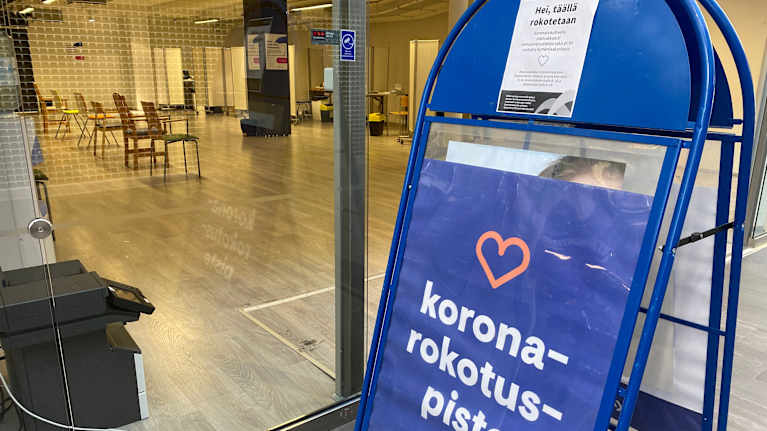 Koronarokotuspiste -teksti a-standissä, jonka takana näkyy tyhjä tila, jossa on tuoleja.