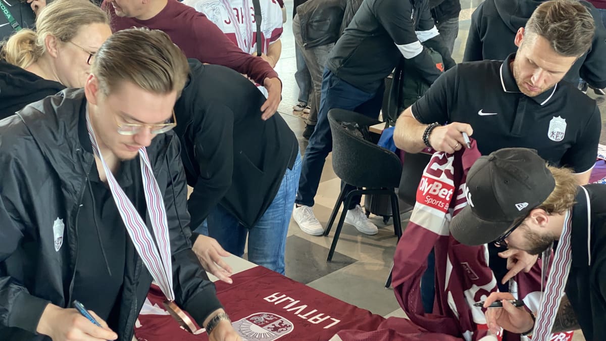 Latvian kiekkojoukkueen kolme pelaajaa signeeraavat punaisia pelipaitoja.