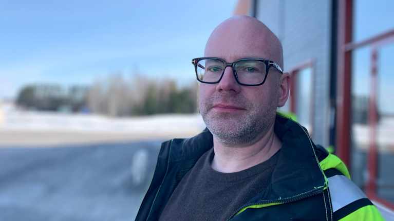 Mansperson i gul reflexjacka står utanför kontorsbyggnad. Ser in i kameran och ler svagt.