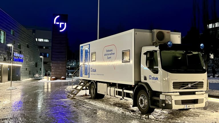  Rekka seisoo talvisella pihamaalla. Rekan kyljessä on Säteilyturvakeskuksen logo STUK, taustalla rakennus jossa on Tampereen yliopiston logo.