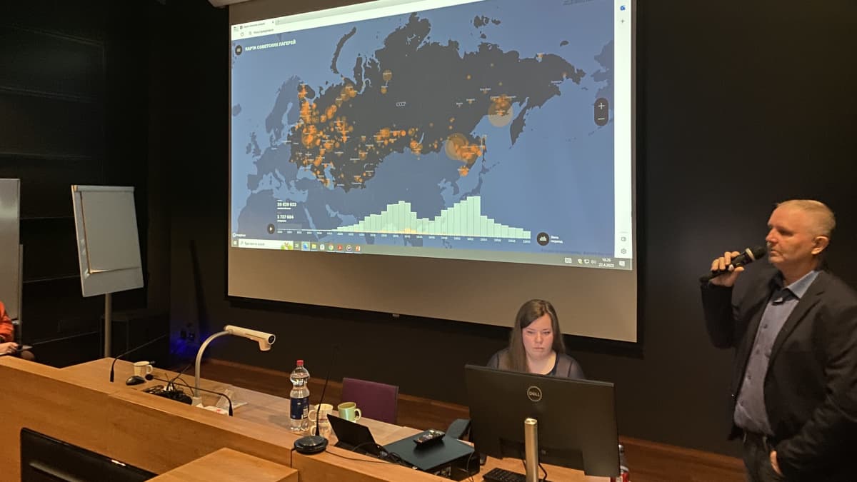 Mies seisoo ja puhuu mikrofoniin, taustalla näkyy isolla näytöllä kartta, jossa on Neuvostoliiton alueella punaisia alueita, ne kuvaavat gulag-vankileirien paikkoja. Professori Puuronen pitämässä esitelmää gulagin historiasta.