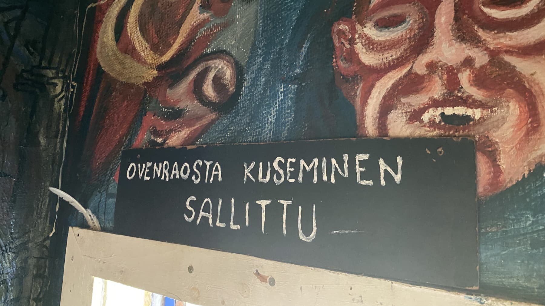 "Ovenraosta kuseminen sallittu" lukee seinässä kyltissä, yläpuolella on myös absurdeja ihmishahmoja.
