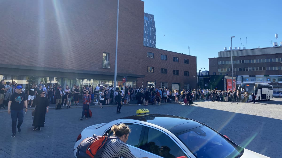 Ihminen kurkottaa taksin ovea. Takana joukko ihmisiä Tampereen rautatieasemalla.