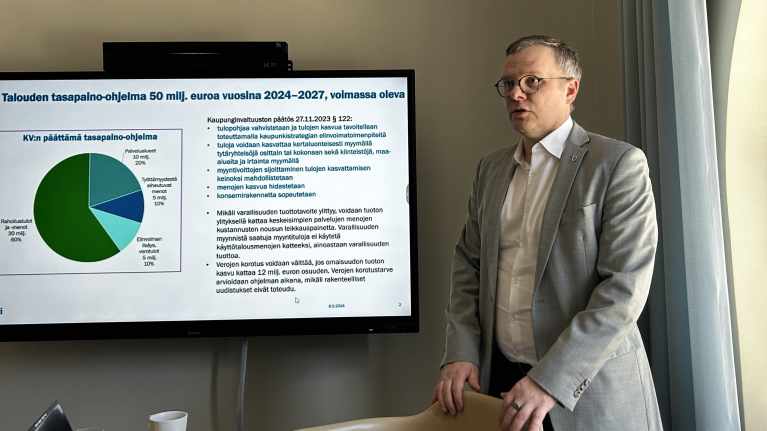 Lahden kaupunginjohtaja Niko Kyynäräinen seisomassa näytön vieressä, ruudulla tekstiä Lahden talouden tasapaino-ohjelmasta.