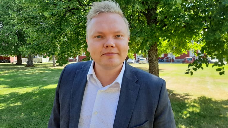 Kulturminister Antti Kurvinen, en blond medelålders man klädd i blå kostym, står under ett träd och tittar in i kameran. 