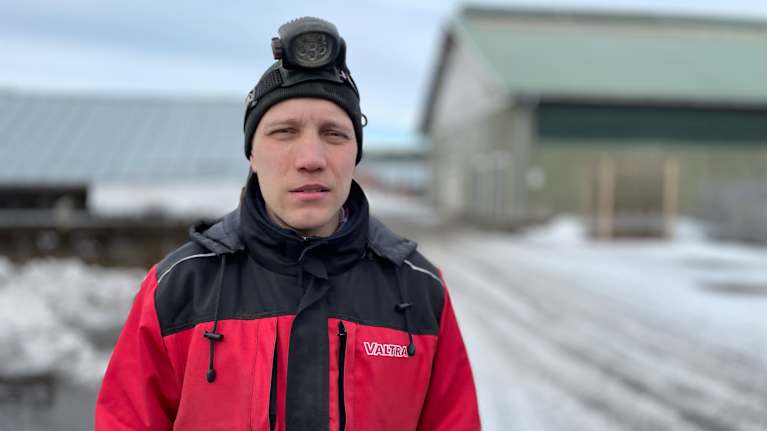 En man i arbetsoverall står ute på en pälsfarm med mössa och pannlampa i snömodden. 