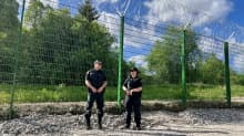 Photo shows two border guards standing in front of a section of fencing, which is about 4 metres tall.