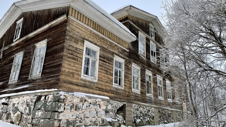 Vanha harmaantunut kartanorakennus takaa kuvattuna talvimaisemassa lumisia lehtipuita oikealla.. 