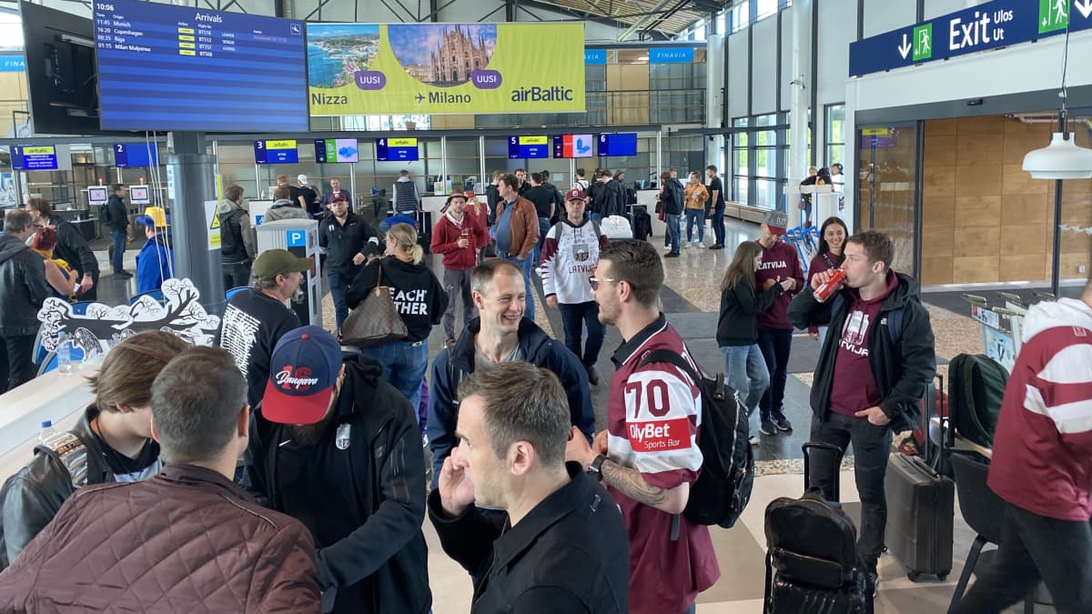 Lentokentällä aulassa seisoo iso joukko ihmisiä. Osa on pukeutunut Latvian punaisiin pelipaitoihin.