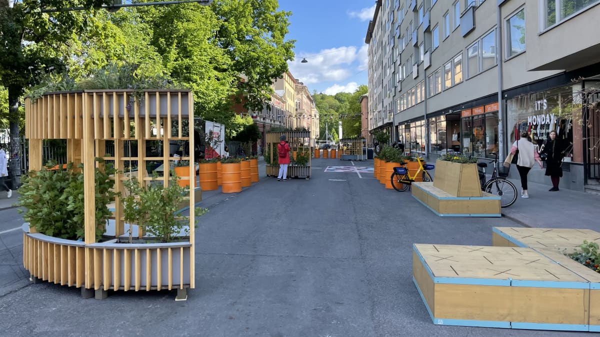 Kristiinankadun kesäkadulle on tuotu puisia istumapaikkoja, oransseja tynnyreitä ja joissa on vihreitä istutuksia. Kadulla on muutamia ihmisiä katsomassa juuri paikalle tuotua rekvisiittaa.