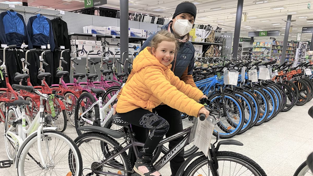 Lapsi (Adessa Kirén) istuu pyörän päällä ja äiti (Heidi Kirén) tukee polkupyörää sivulta. Takana eri värisiä lastenpyöriä jonoissa.