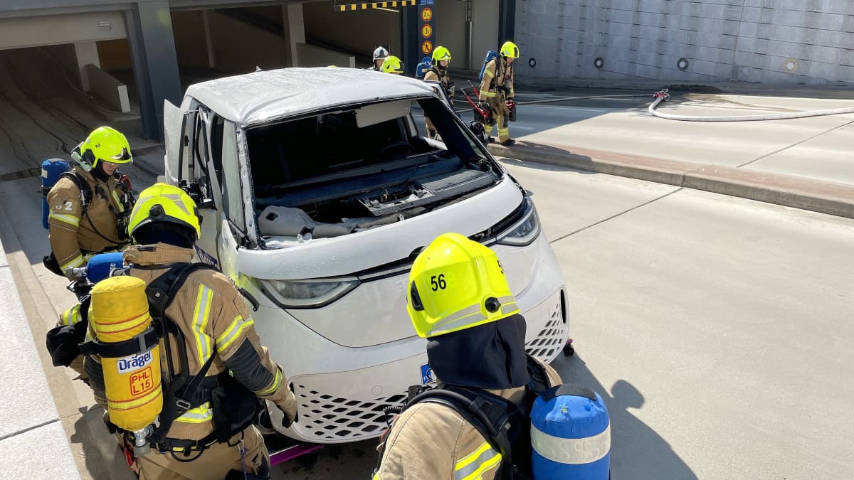 Etualalla kolme palomiestä tutkailee palanutta, valkoista sähköautoa. Auton takana neljä palomiestä. Autoa hinataan parkkihallista tielle.