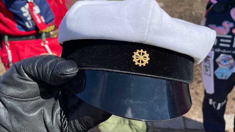 Opiskelija pitää kädessään ylioppilaslakkia. Takana näkyy opiskelijahaalareita ja vappuilmapiiriä.