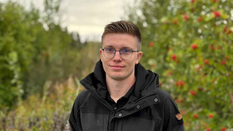 En person med glasögon och svart jacka står utomhus framför grönska och rönnbärsträd. Ingen synlig text.
