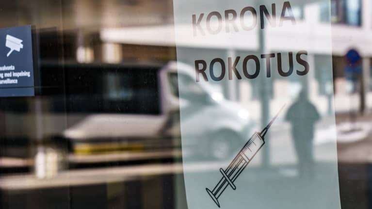 Koronarokotuspaikan lasiovesta heijastuu  ihmisen hahmo. Ikkunassa lappu, jossa lukee koronarokotus.