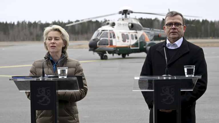 Ursula von der Leyen och Petteri Orpo i vinterkläder står vid talarpodium på en flygplats och pratar. I bakgrunden en helikopter.