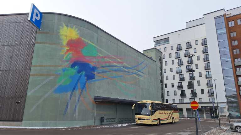 Kangasalan pysäköintitalo Kuohunparkki. Vihertävässä seinässä on värikäs, abstrakti seinämaalaus. Keltainen bussi on pysäköitynä rakennuksen eteen.